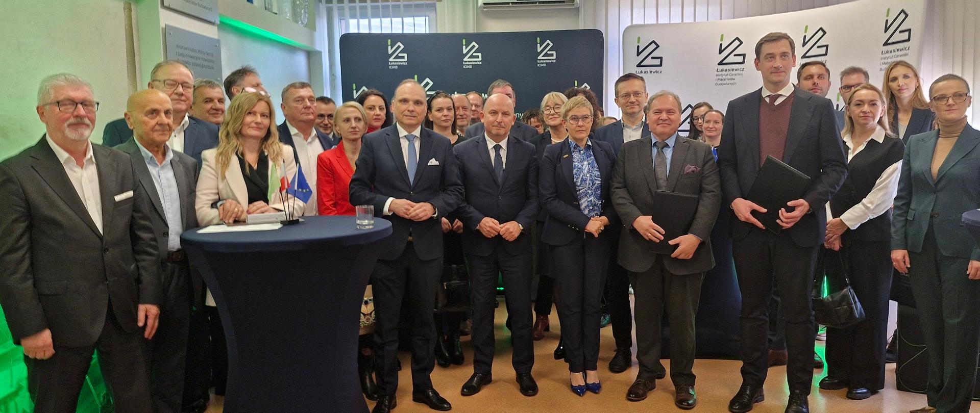 Zdjęcie zbiorowe, na sali stoi grupa około 20 osób, pośrodku grupy wiceminister Mrówczyńska, za nimi rozstawione ścianki z napisem Łukasiewicz, w tle zasłonięte poziomymi żaluzjami okna.