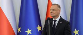 Prezydent Duda stoi i mówi do mikrofonu na stojaku, za nim pod ścianą flagi Polski i UE.