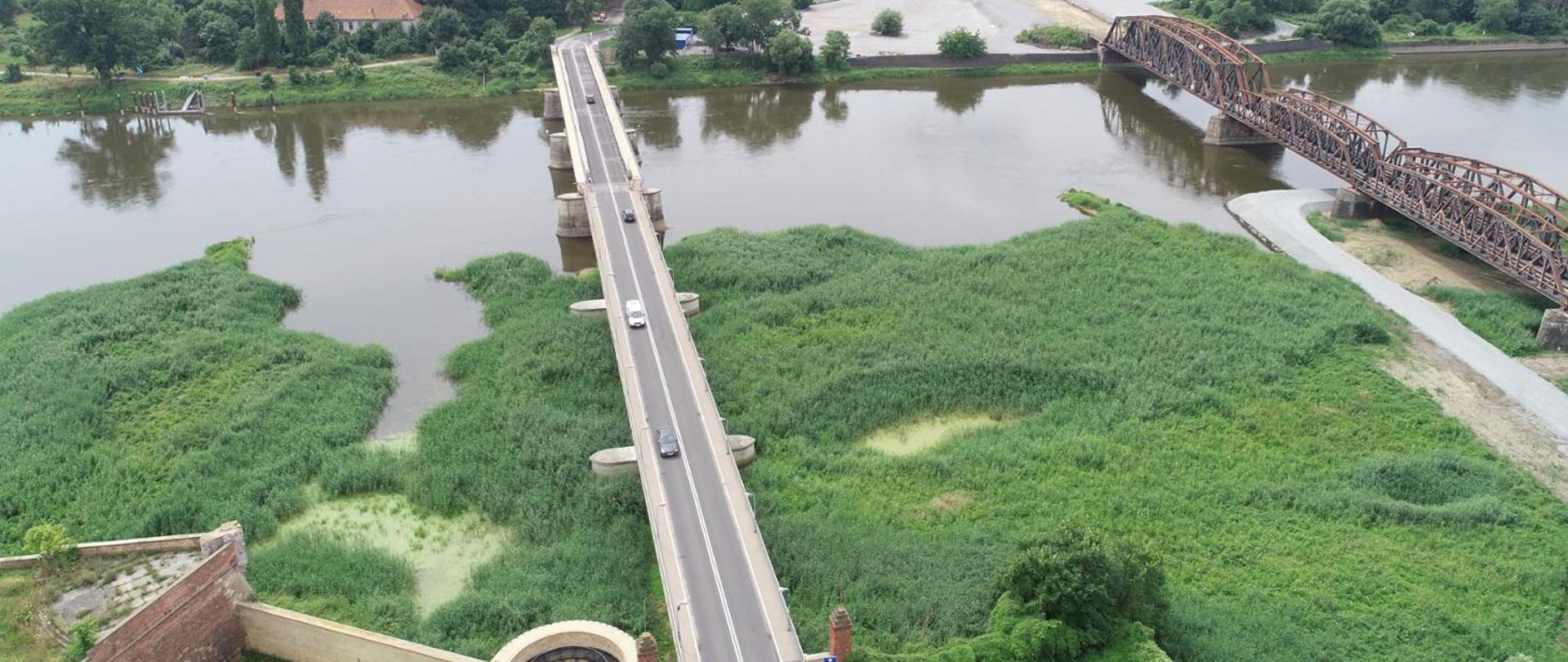 Most drogowy rozpięty nad nurtem rzeki w towarzystwie mostu kolejowego. Oba otoczone zielenią, widoczne nieliczne zabudowania oraz fragment zabytkowej twierdzy.