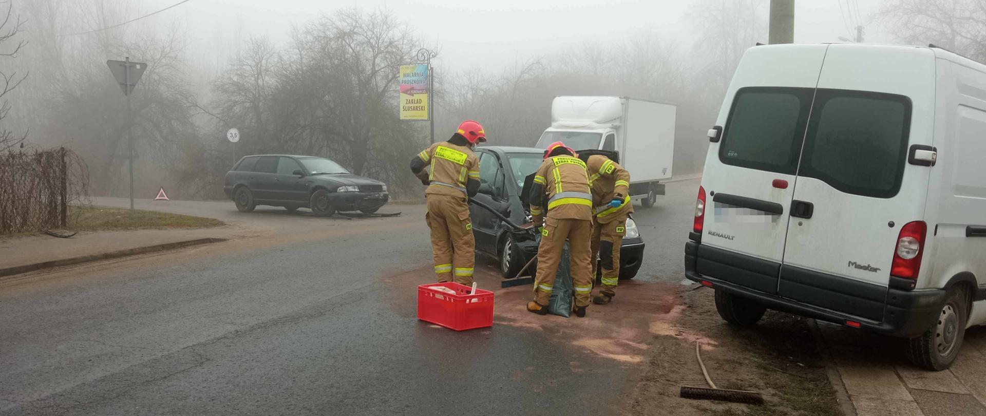 Zdjęcie zrobione na ul. Portowej w miejscu kolizji trzech pojazdów. Na zdjęciu widać samochód dostawczy i dwa samochody osobowe biorące udział w zdarzeniu. Przy samochodzie osobowym i dostawczym trzech strażaków porządkuje jezdnię z rozlanych płynów eksploatacyjnych. 