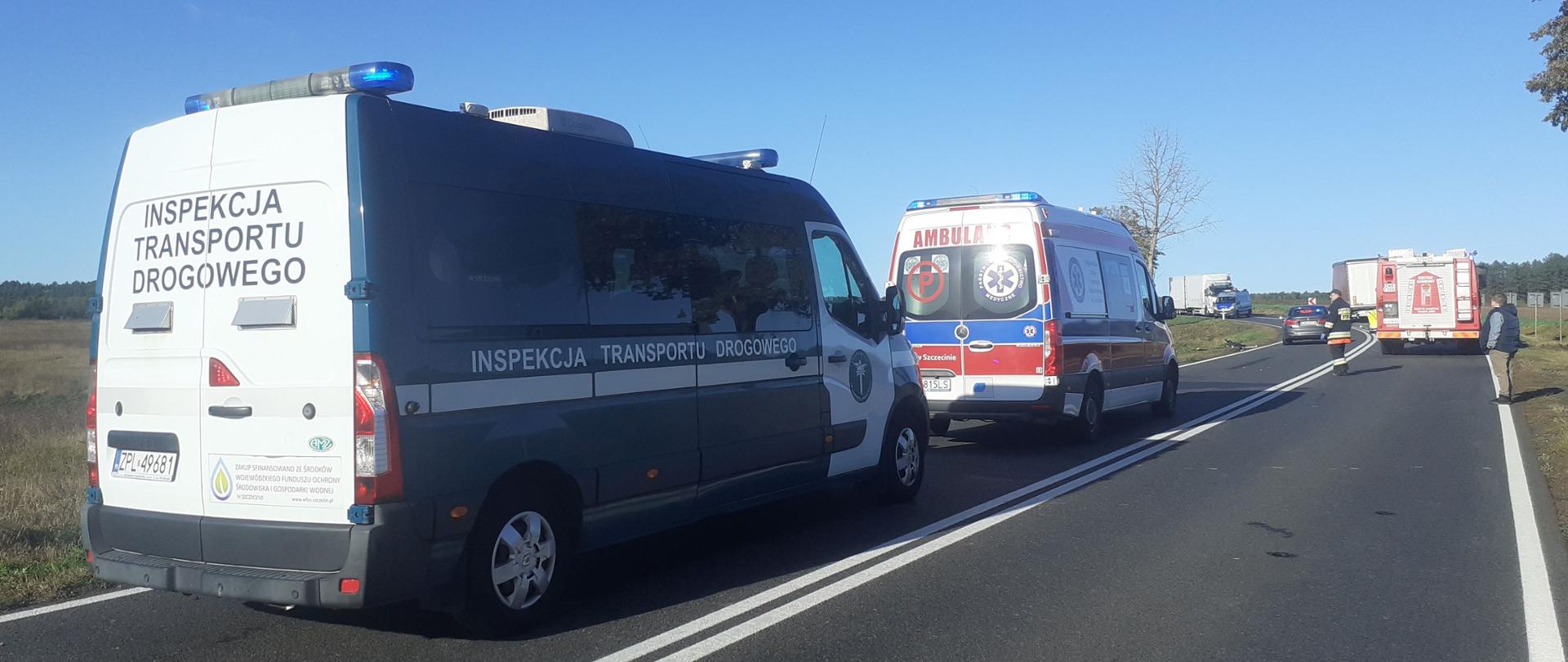 Radiowóz Inspektoratu Transportu Drogowego stojący za karetką pogotowia ratunkowego, w oddali wóz ochotniczej straży pożarnej, nieoznakowany radiowóz policji oraz zniszczony pojazd ciężarowy.