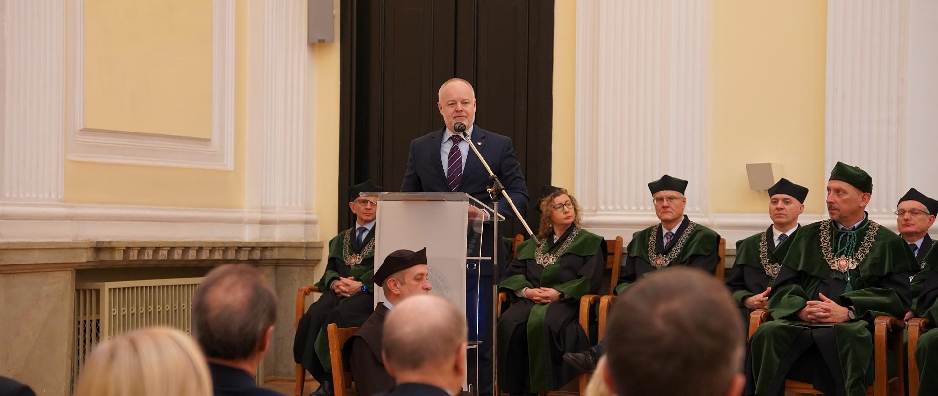 Wiceminister Gzik stoi za mównicą i mówi do mikrofonu, za nim na krzesłach siedzi kilka osób w zielonych ceremonialnych strojach, przed nim wypełniona siedzącymi w rzędach krzeseł ludźmi.