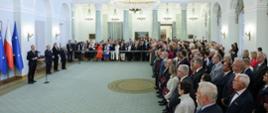 Widok z boku na dużą salę, z lewej prezydent Duda stoi i mówi do mikrofonu, za nim flagi Polski i UE, przed nim widać dużo ludzi.