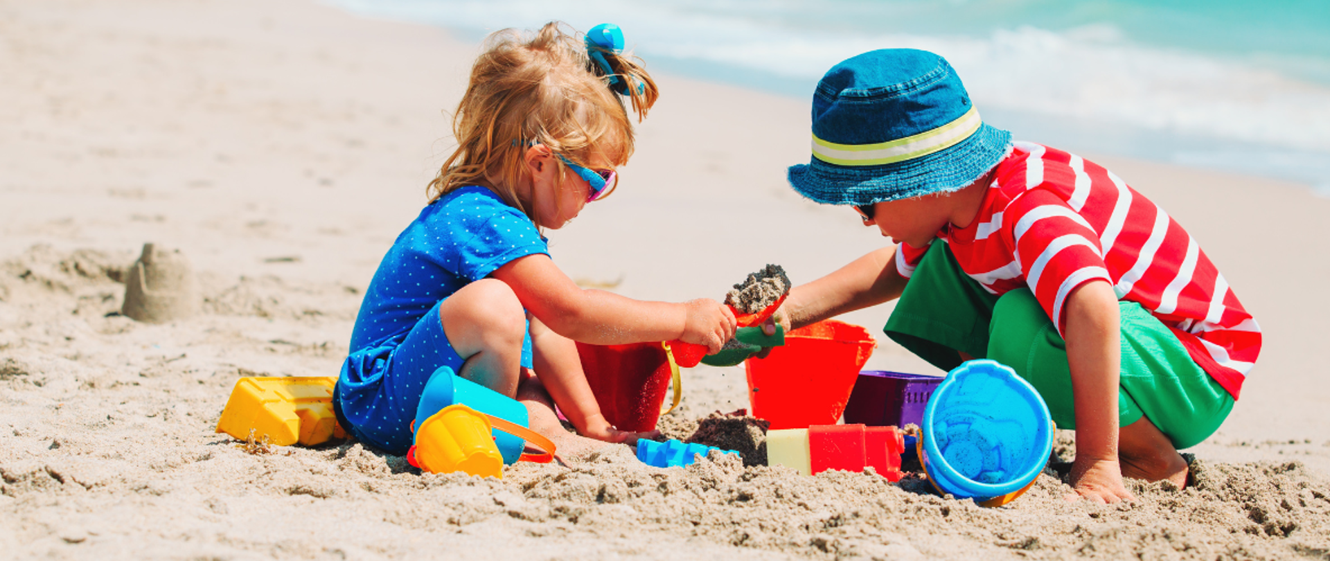 dzieci bawią się na piaszczystej plaży, z lewej mała dziewczynka sypie piasek łopatką do kolorowych foremek, po prawej starszy chłopiec w kapeluszu trzyma foremkę, na około kolorowe zabawki plażowe do piasku 