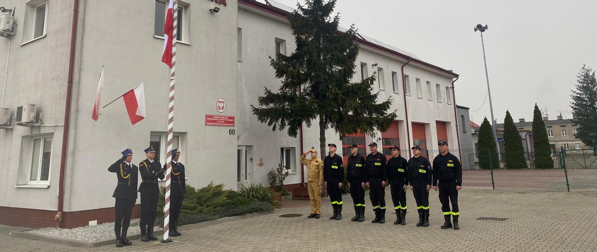 Zdjęcie przedstawia poczet flagowy oraz strażaków stojących w szyku podczas wciągnięcia na masz flagi państwowej oraz odśpiewaniem hymnu państwowego 