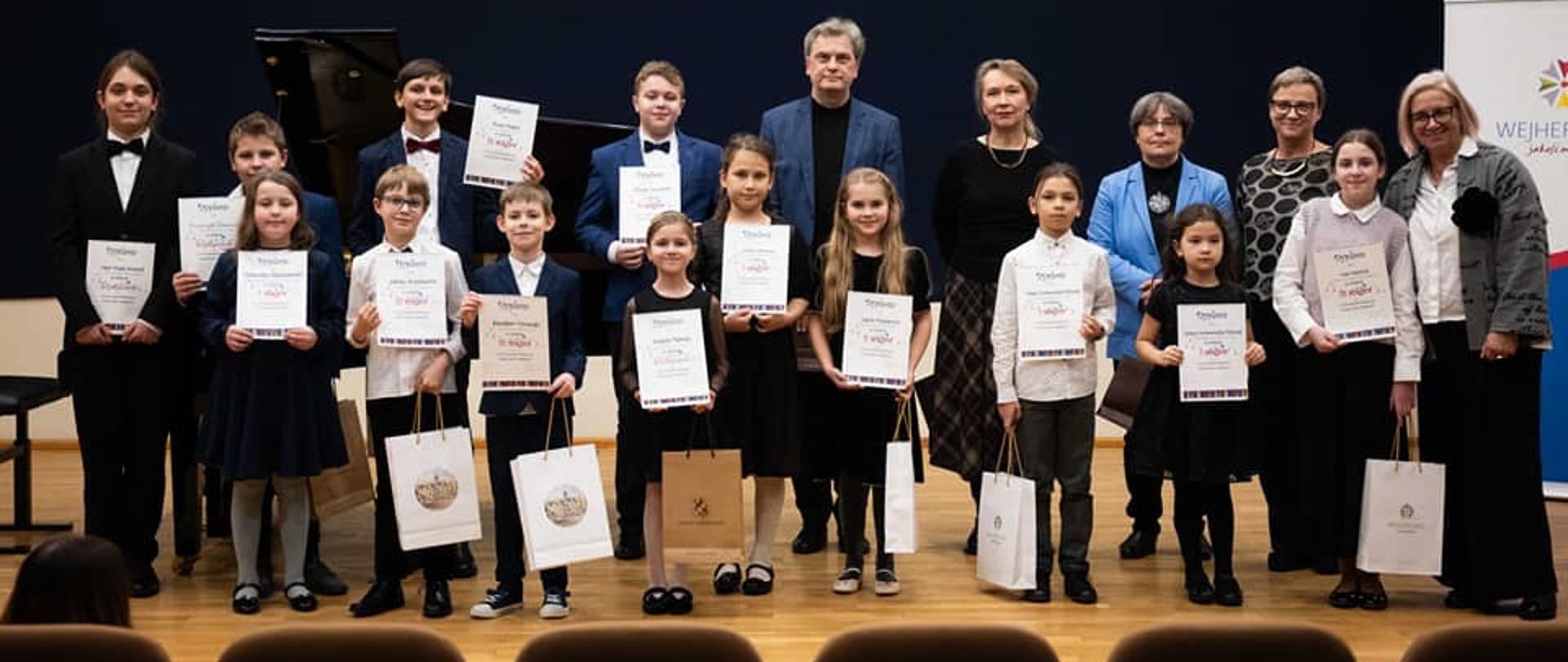 Na estradzie pozują do zdjęcia laureaci konkursu wraz z jurorami i nauczycielami gry na fortepianie. Każde dziecko eksponuje dyplom. W tle na ścianie nad granatową podłużną płachtą napis: "Państwowa Szkoła Muzyczna I st. im. Fryderyka Chopina w Wejherowie".