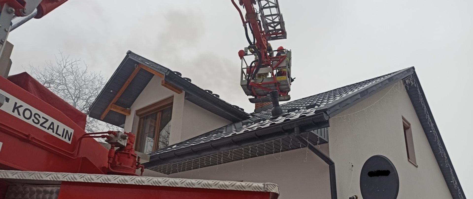 Na zdjęciu pojazd pożarniczy specjalny-podnośnik, w koszu podnośnika strażacy prowadzący działania gaśnicze przewodu kominowego, w tle zdjęcia budynek mieszkalny
