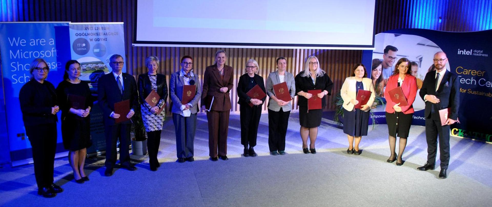 Na scenie stoi grupa osób wśród nich Minister Barbara Nowacka. W tle na dużym ekranie napis powodzenia