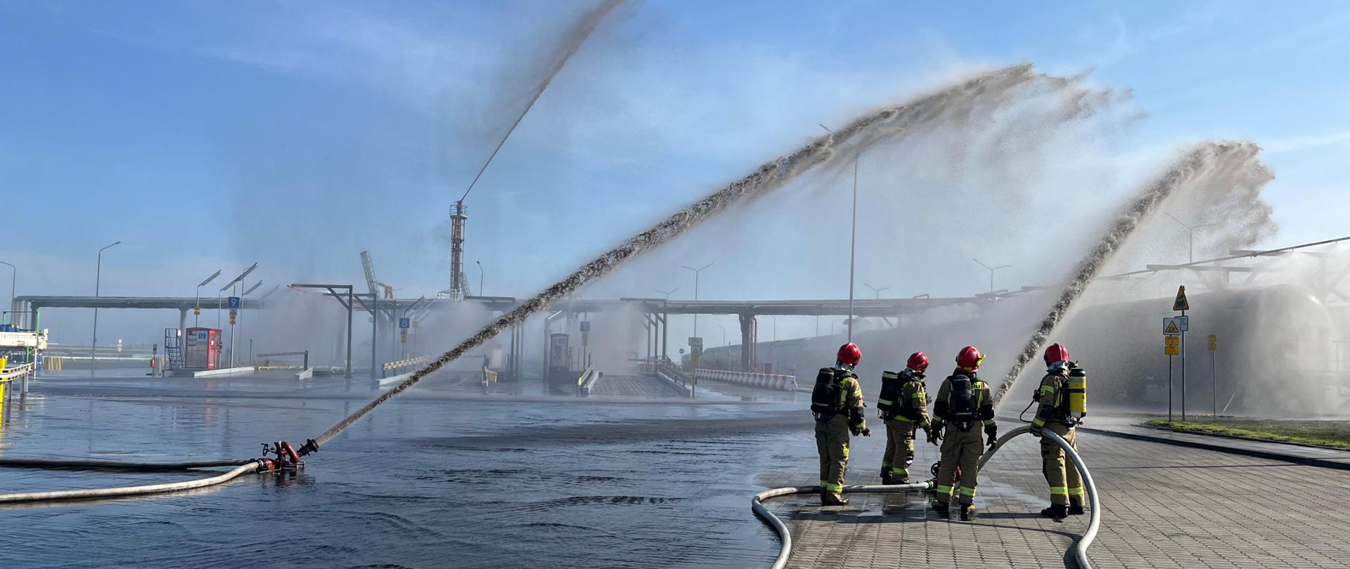 Widok symulowanego pożaru cysterny z LPG. Strażacy podają wodę z działek na cysternę.