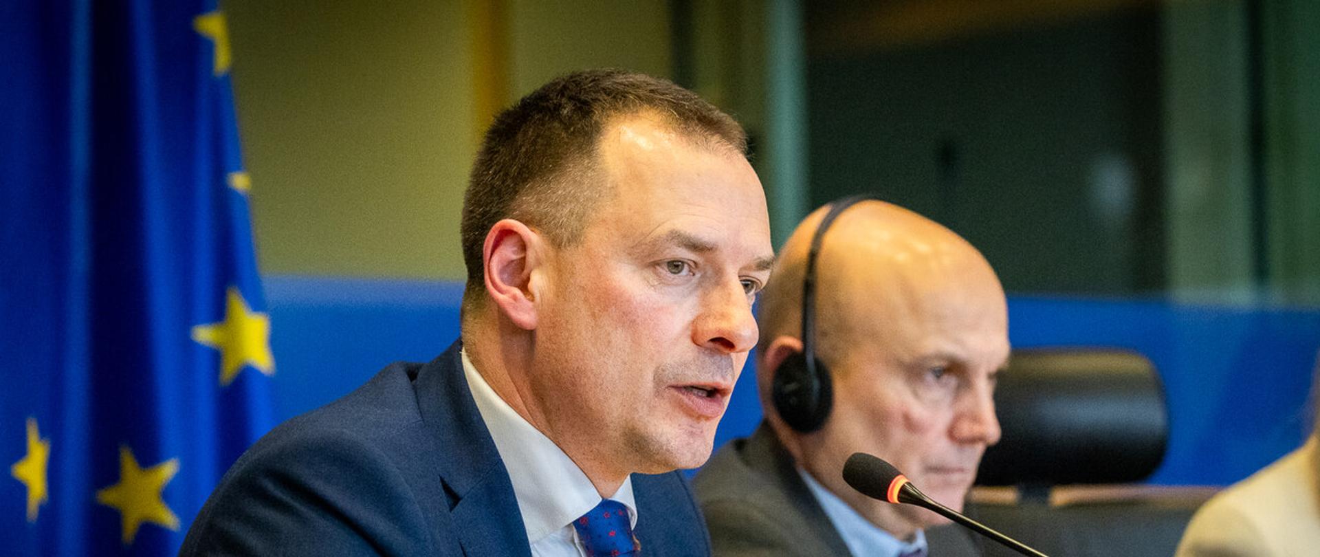 Piotr Borys przed Komisją Transportu i Turystyki przed mikrofonem na tle flagi Unii Europejskiej