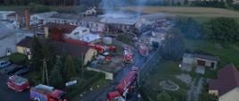 Zdjęcie przedstawia pożar zakładu do produkcji papieru. Zdjęcie z lotu ptaka. Na zdjęciu widać unoszący się dym nad jednym z budynków. Ponadto widać samochody strażackie i strażaków. 