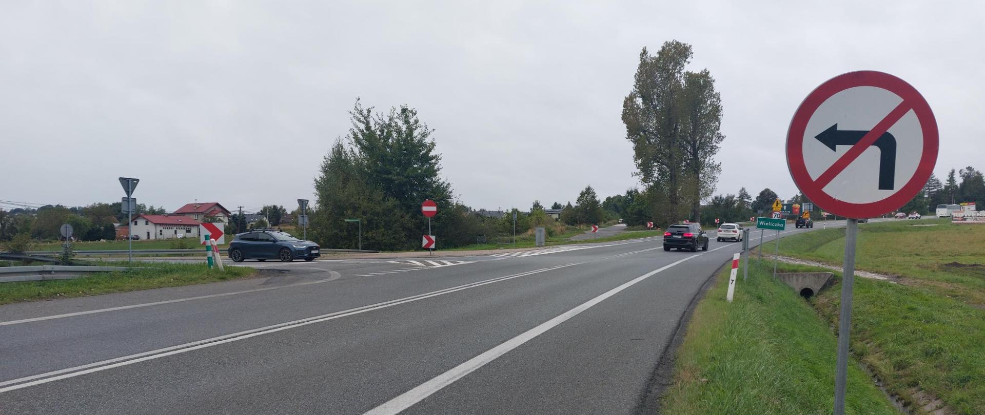 Zdjęcie przedstawia skrzyżowanie. Na pierwszym planie po prawej stronie znajduje się znak z zakazem skrętu w lewo. Widać tablicę z nazwą miejscowości - Wieliczka