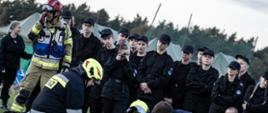 Stadion w Miejskiej Górce. Trwa pokaz akcji ratowniczej w związku z zasymulowanym wypadkiem drogowym. Na tle klasy mundurowej strażacy udzielaja kwalifikowanej pierwszej pomocy ofierze wypadku. Z lewej strony stoi kierujący działaniem ratowniczym. W tle namioty i drzewa.