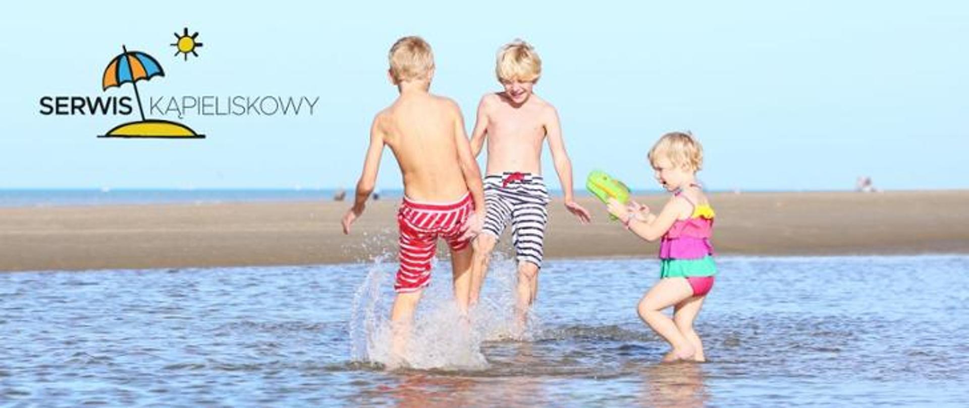 Dzieci w strojach kąpielowych bawiące się w wodzie, najprawdopodobniej w morzu. Na dalszym tle widać piaszczystą plażę.