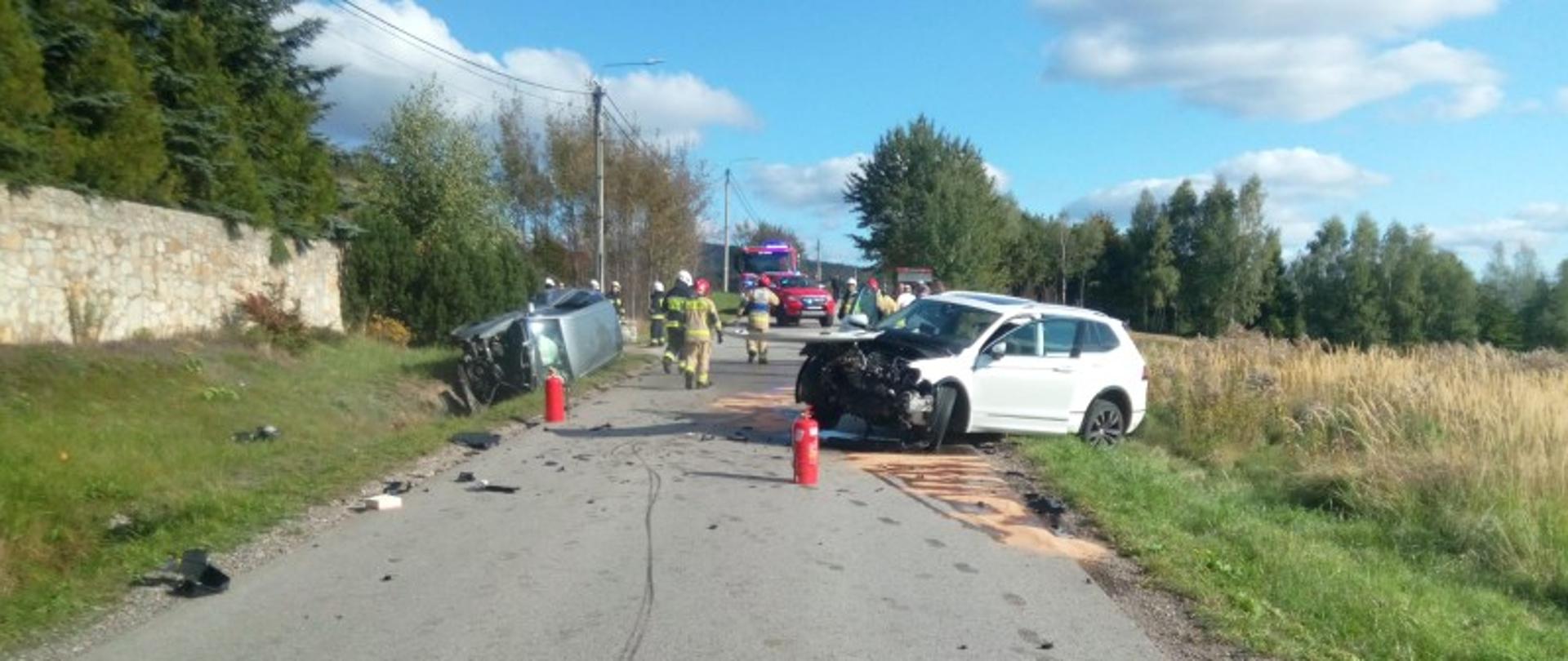 Zdjęcie przedstawia drogę gminną i zniszczone w wyniku zderzenia dwa samochody znajdujące się po dwóch stronach jezdni.