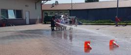Dzieci wraz ze strażakiem PSP lejące wodę z prądownicy do pachołków. W tle budynki