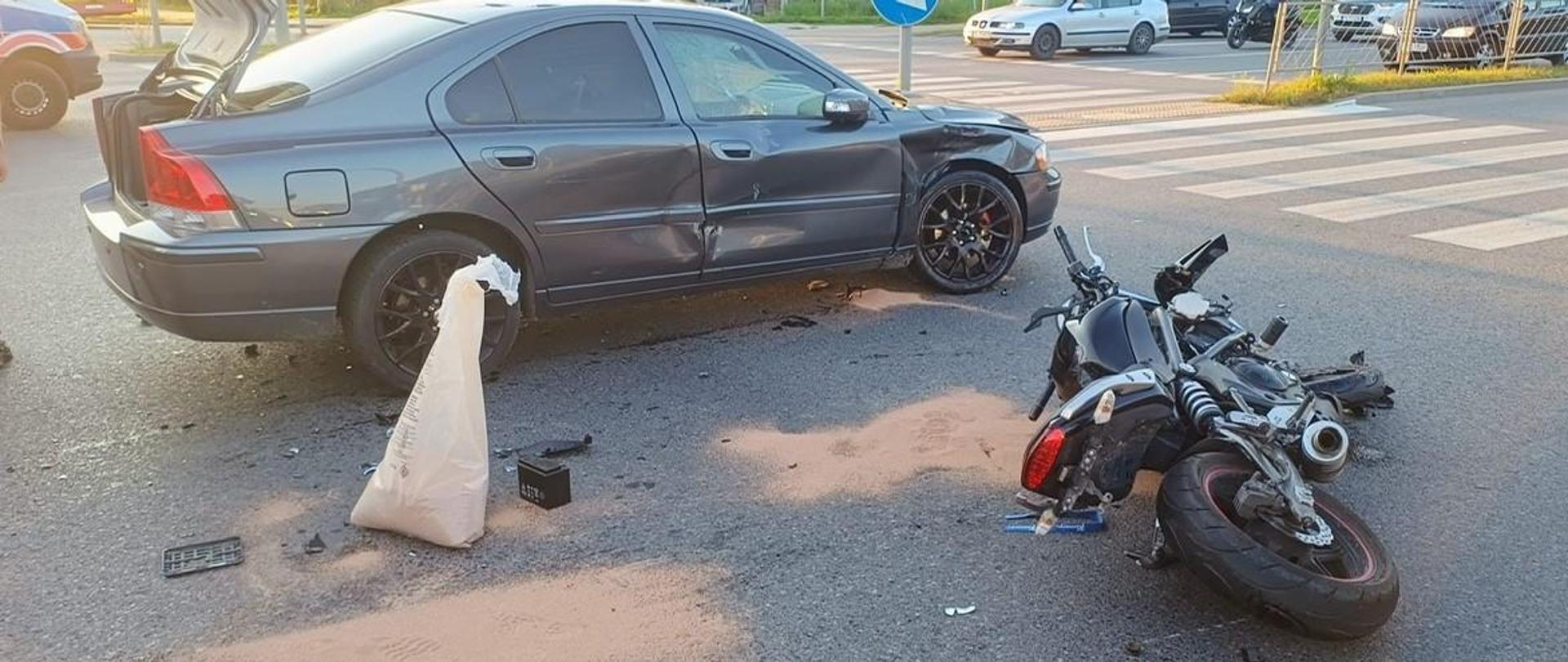 Zdjęcie przedstawia samochód osobowy i przewrócony na jezdnię motocykl. Widać plamy płynów eksploatacyjnych posypane sorbentem.