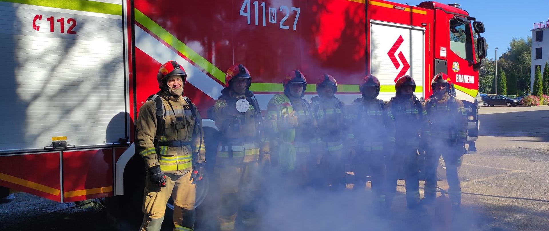 Na zdjęciu na tle dużego samochodu strażackiego stoi siedmiu strażaków w mundurach bojowych i czerwonych hełmach, unosi się dym.