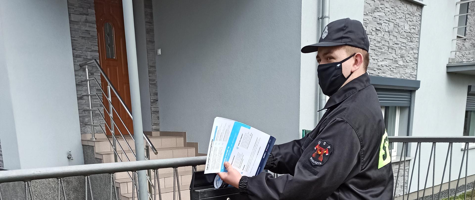 Zdjęcie przedstawiające Strażaka OSP umieszczającego w skrzynce pocztowej ulotkę na temat akcji informacyjnej #SZCZEPIMY SIĘ