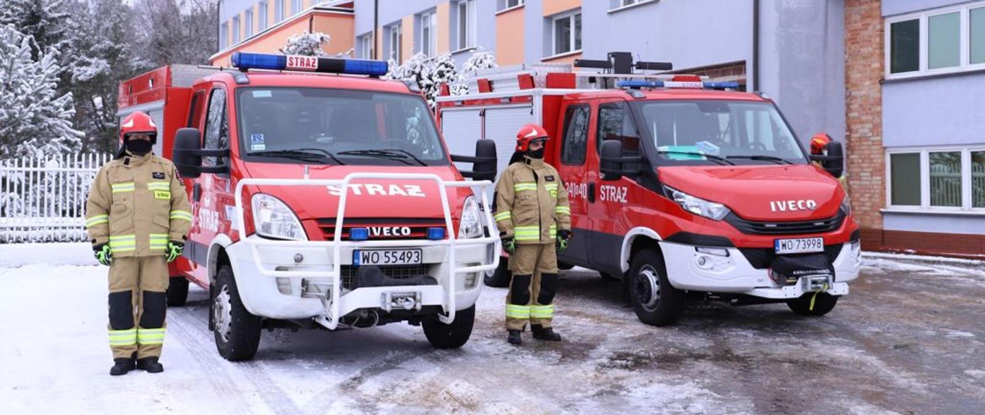 Na zdjęciu dwa samochody straży pożarnej, przy których swoi dwóch strażaków w mundurach.