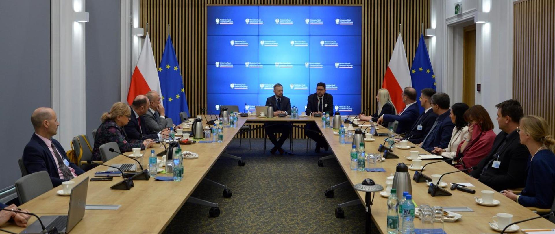 Zdjęcie od tyłu sali, za ustawionymi w czworokąt stołami siedzi dużo ludzi, u szczytu stołu wiceminister Szeptycki, za nim na ścianie wielki ekran, po obu jego stronach flagi Polski i UE.