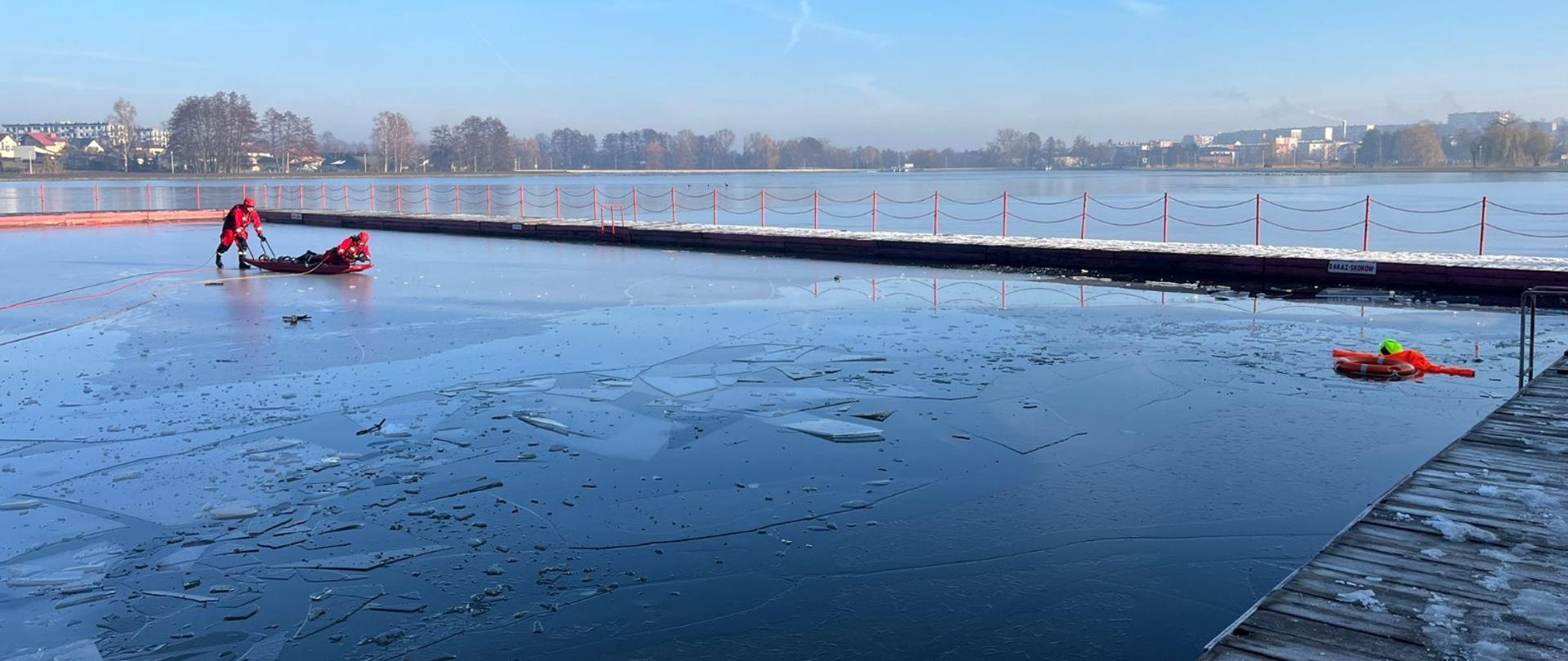 Na zbiorniku wodnym dwóch strażaków przy użyciu sani lodowych woda-lód przemieszcza się do pozorowanego poszkodowanego, którym jest manekin. Jeden ze strażaków znajduje się na saniach, a drugi pcha te sanie.