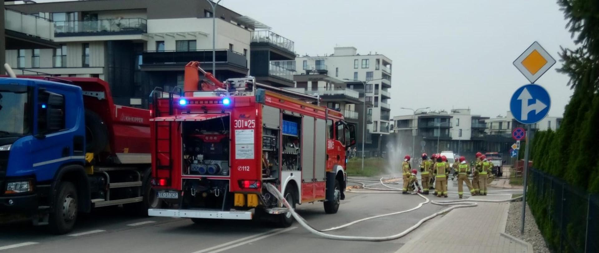 W centralnym punkcie stoi zaparkowany samochód gaśniczy z podpiętymi wężami zasilającymi kurtyny wodne. W głębi, po prawej stronie, stoi grupa strażaków przed działającymi kurtynami. 