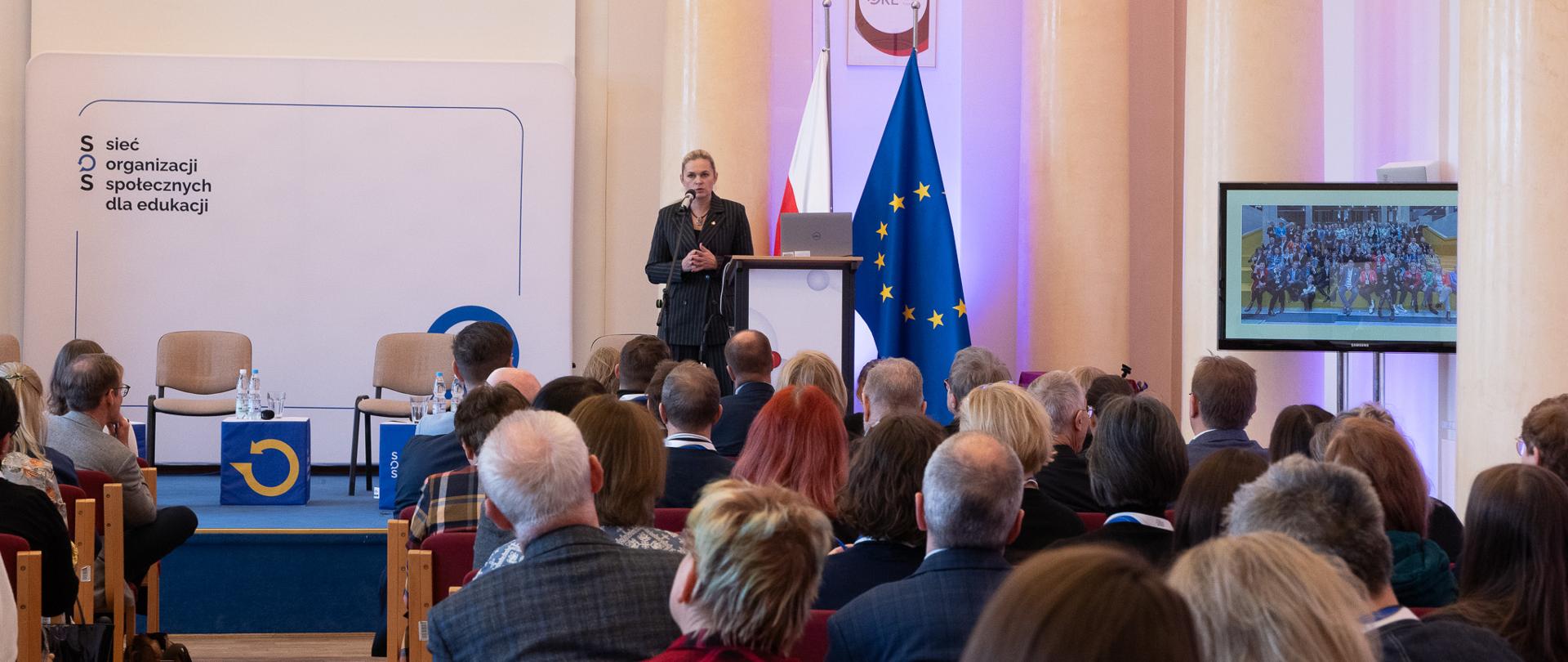 Widok z tyłu na salę, na ustawionych w rzędy krzesłach siedzą ludzie. Z przodu na podwyższeniu stoi minister Nowacka obok pulpitu i przemawia do mikrofonu. Na scenie ustawione widać dwa krzesła i kwadratowe stoliki na których stoją butelki z wodą