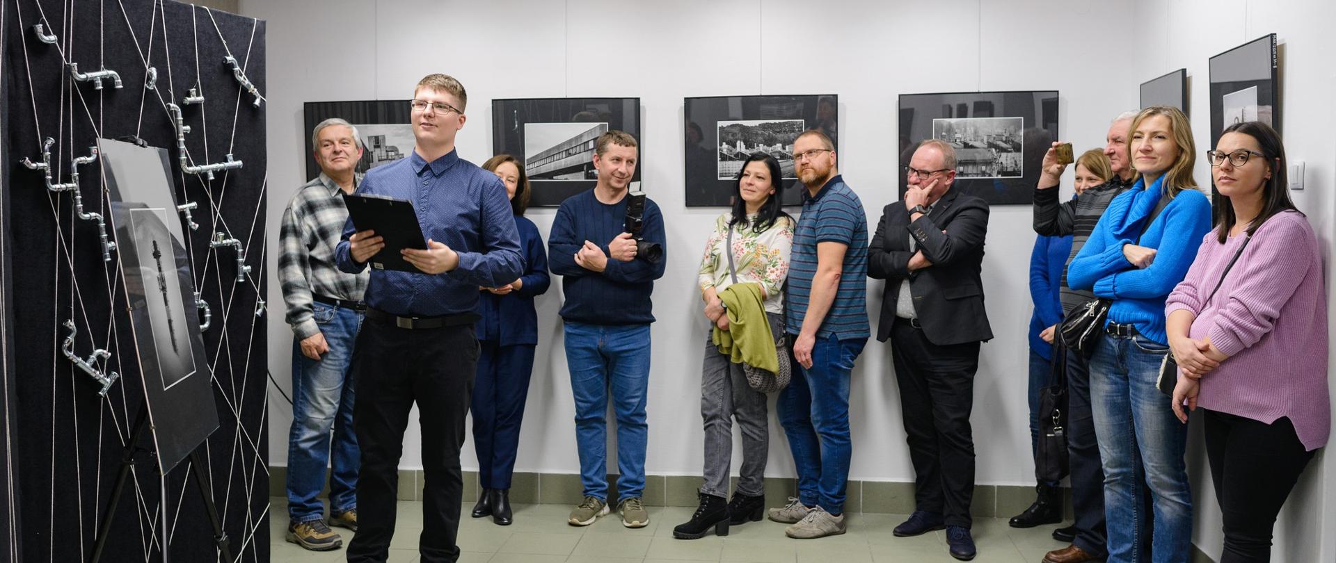 Na zdjęciu stoją osoby które przyszły na wernisaż wystawy Michała Niemca. Stoją na tle zdjęć które są zawieszone na białej ścianie. Po lewej stronie stoi autor wystawy a obok niego czarny parawan na którym są zaczepione srebrne rurki. 