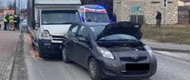 Zdjęcie przedstawia uszkodzony pojazd Toyota, na którego tył najechał pojazd dostawczy. Za pojazdami widać funkcjonariuszy PSP i Policji