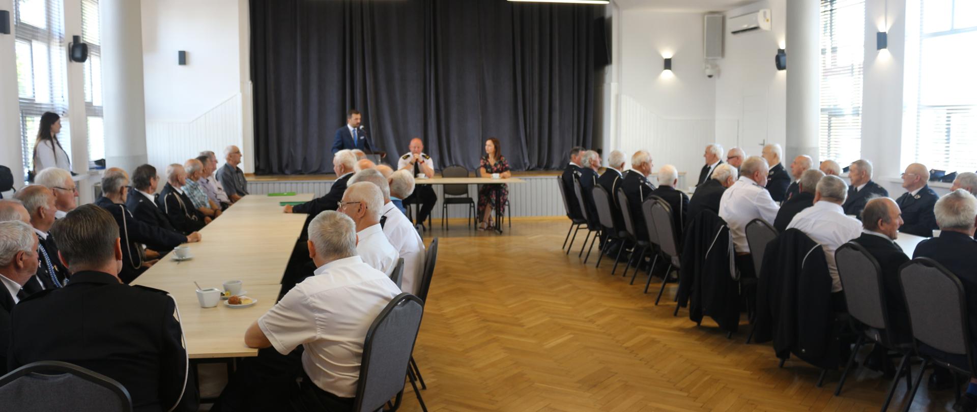 Druhowie siedzą przy stołach w sali centrum kultury. Z mównicy przemawia burmistrz miasta Grabowa nad Prosną.