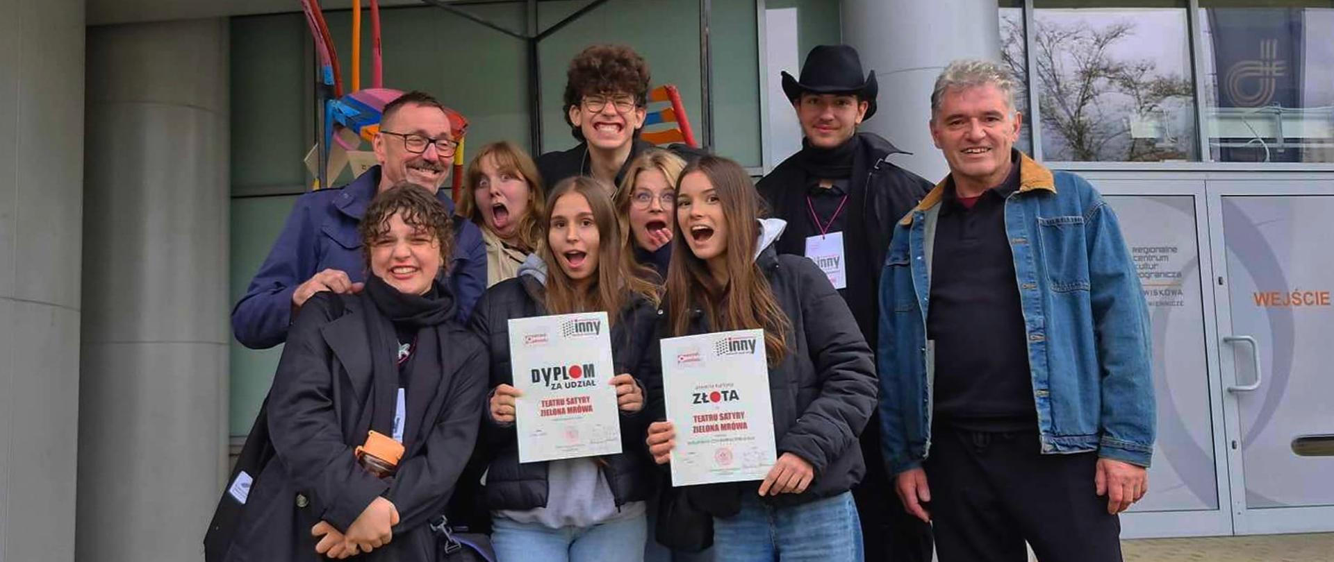 Na zdjęciu stoi przed budynkiem grupa czterech mężczyzn i czterech dziewcząt. Dwie dziewczyny trzymają dyplomy. Wszyscy uśmiechają się i robią śmieszne miny.