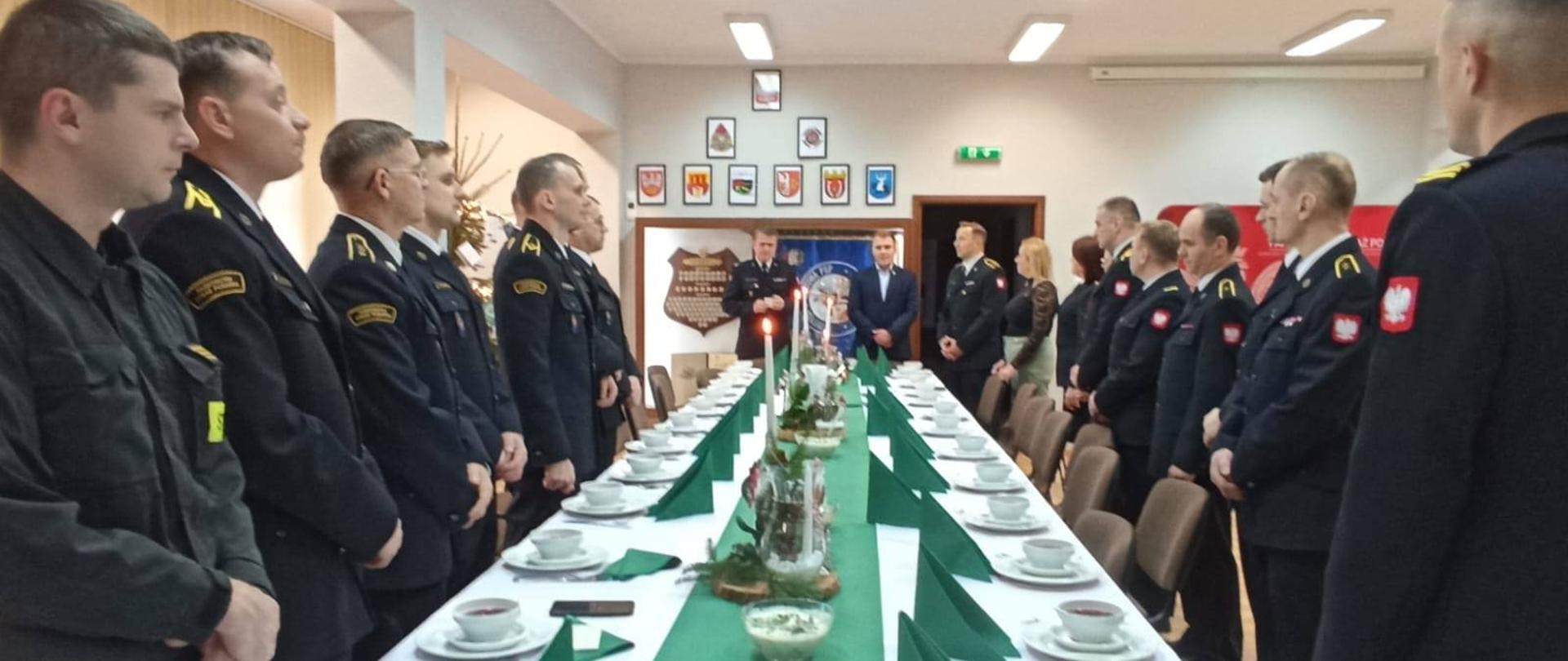 Zdjęcie przedstawia stojących strażaków podczas spotkania wigilijnego.
W tle pomieszczenie świetlicy.
