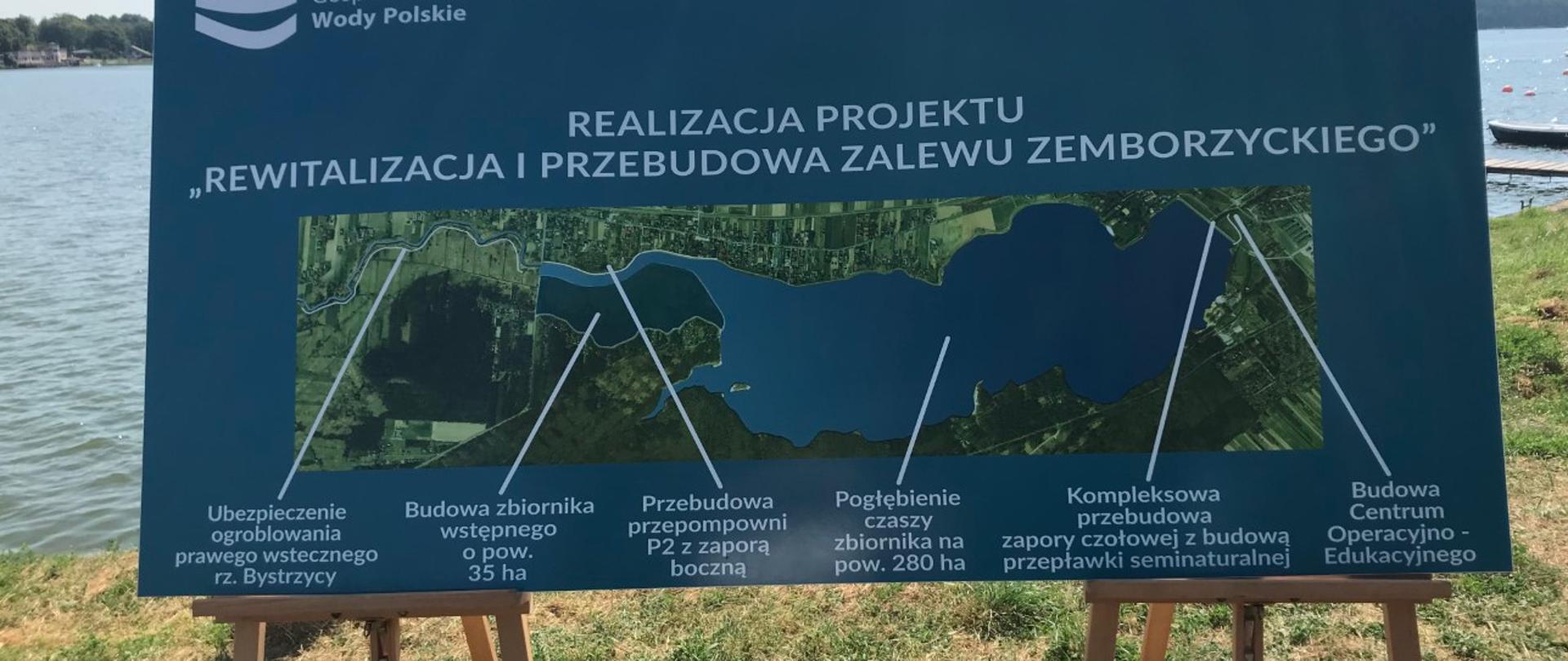 Dwie sztalugi malarskie ustawione na trawie. W tle zbiornik wodny. Na sztalugach oparty plakat przedstawiający mapę Zalewu Zemborzyckiego. 