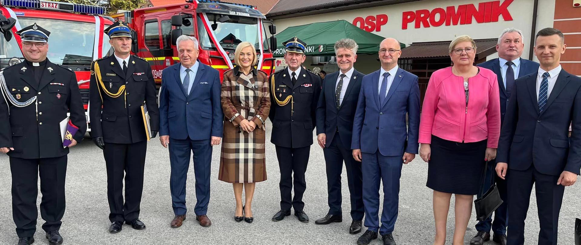 Zdjęcie przedstawia zaproszonych gości na uroczystości przekazania i poświęcenia nowego samochodu na tle remizy Ochotniczej Straży Pożarnej w Promniku oraz dwóch wozów pożarniczych: nowego dla OSP Promnik i przekazywanego dla OSP Oblęgorek.