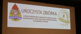 Okolicznościowy slajd tytułowy wyświetlany na ekranie w auli Szkoły. Napis – uroczysta zbiórka z okazji rozpoczęcia kwalifikacyjnego kursu zawodowego oraz szkolenia podstawowego w zawodzie strażak 