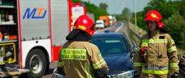Na zdjęciu widocznych jest dwóch strażaków. za nimi po lewej stronie znajduje się samochód ratowniczo gaśniczy.