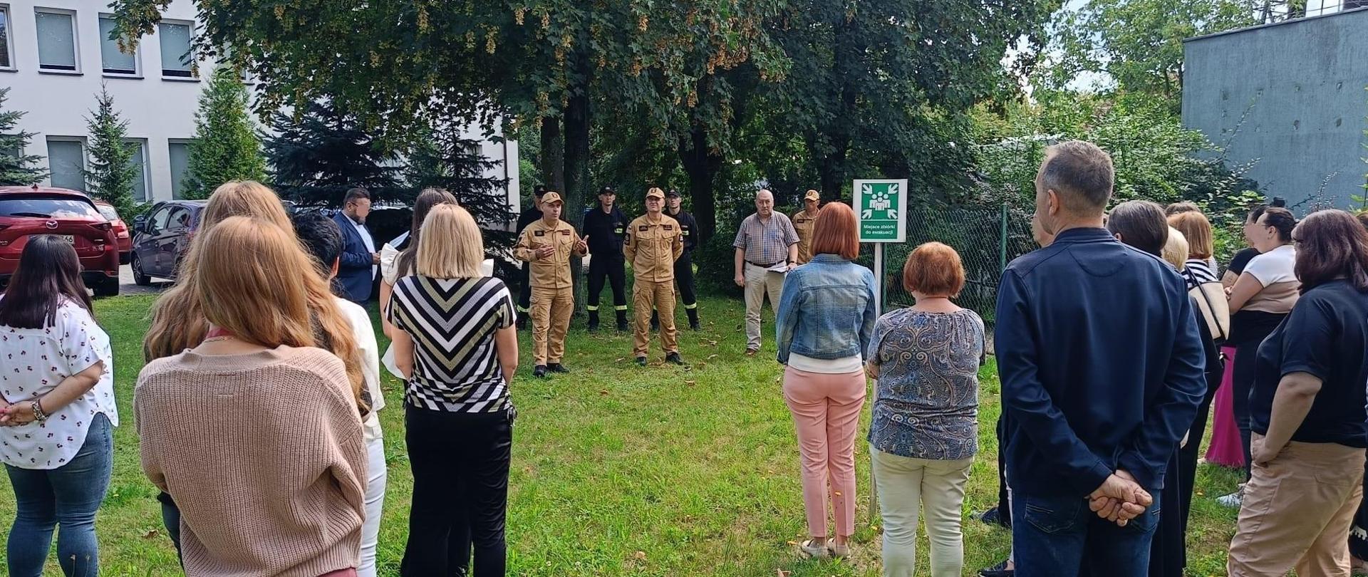 Miejsce zbiórki na zewnątrz budynku, na którym stoi grupa kobiet i mężczyzn, na wprost nich stoi czterech strażaków, jeden z nich ubrany w żółty mundur Państwowej Straży Pożarnej zwraca się do cywilów