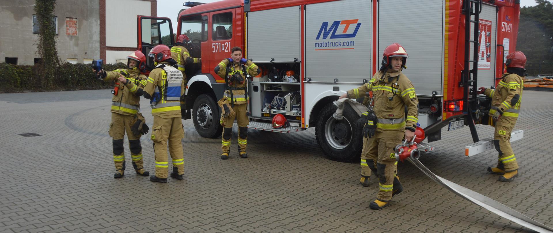 Na placu manewrowym stoi wóz strażacki, wokół którego znajdują się strażacy przygotowujący się do akcji. W tle budynek.