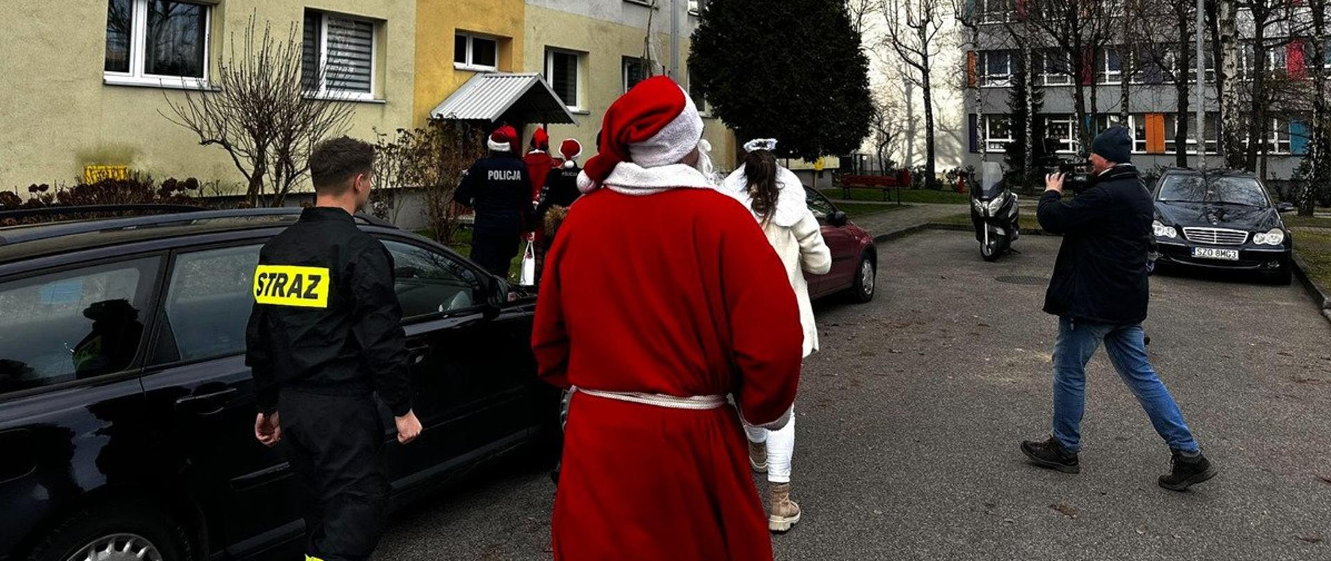 Grupa ludzi idzie w kierunku budynku wielorodzinnego wśród nich strażak, policjant oraz przebrani za Świętego Mikołaja i aniołki. Obok zaparkowane samochody. 