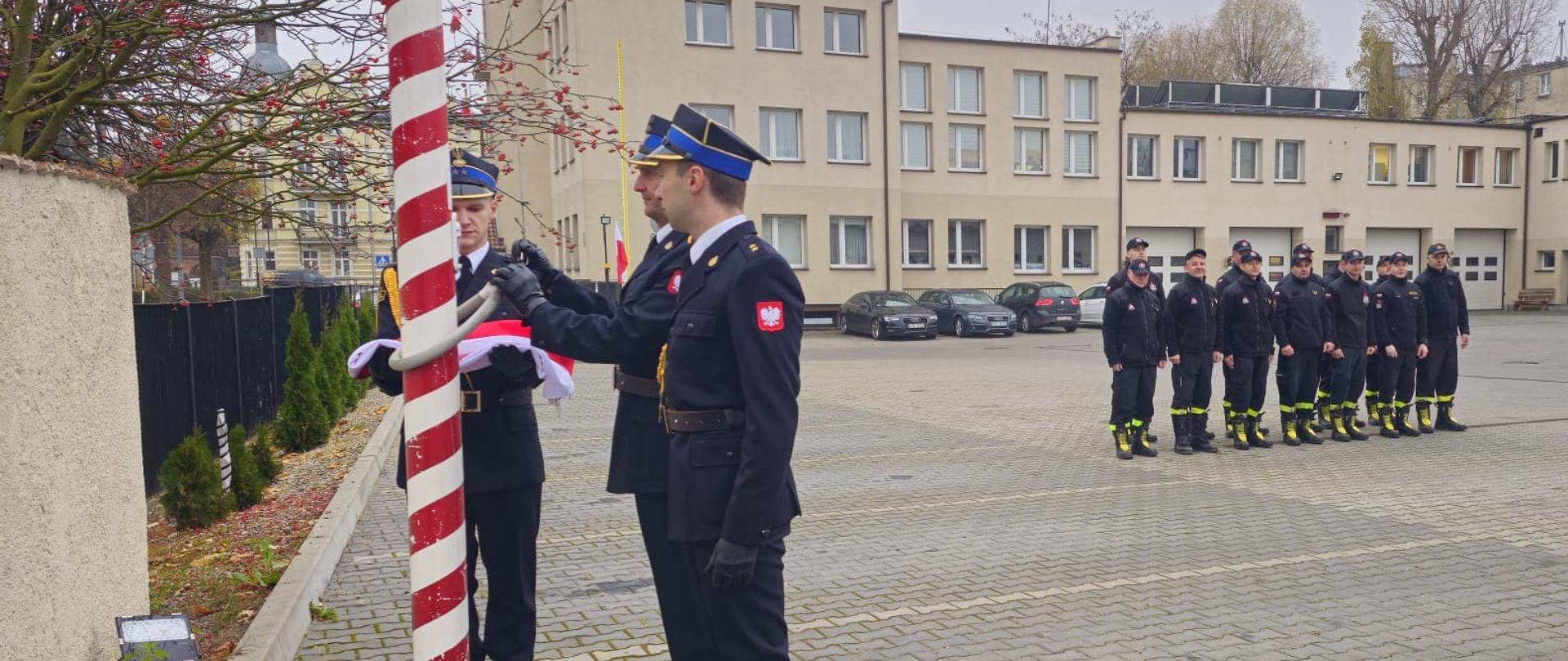 Plac KP PSP Tczew, poczet flagowy z flagą, obok ustawiony pododdział strażaków. W tle budynek komendy i ratusz.