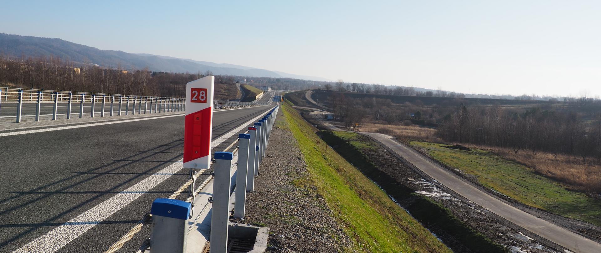 Widok z poziomu ziemi na drogę. Okres wiosenny, świeci słońce, drzewa jeszcze bez liści ale powoli zieleni się trawa. Na pierwszym planie widać słupek pikietażowy z numerem 28 informującym jaka to droga. Widać drogę z namalowanym oznakowaniem poziomym, bariery energochłonne. Po prawej stronie jest rów i jezdnia serwisowa. W tle widać wzgórza. 