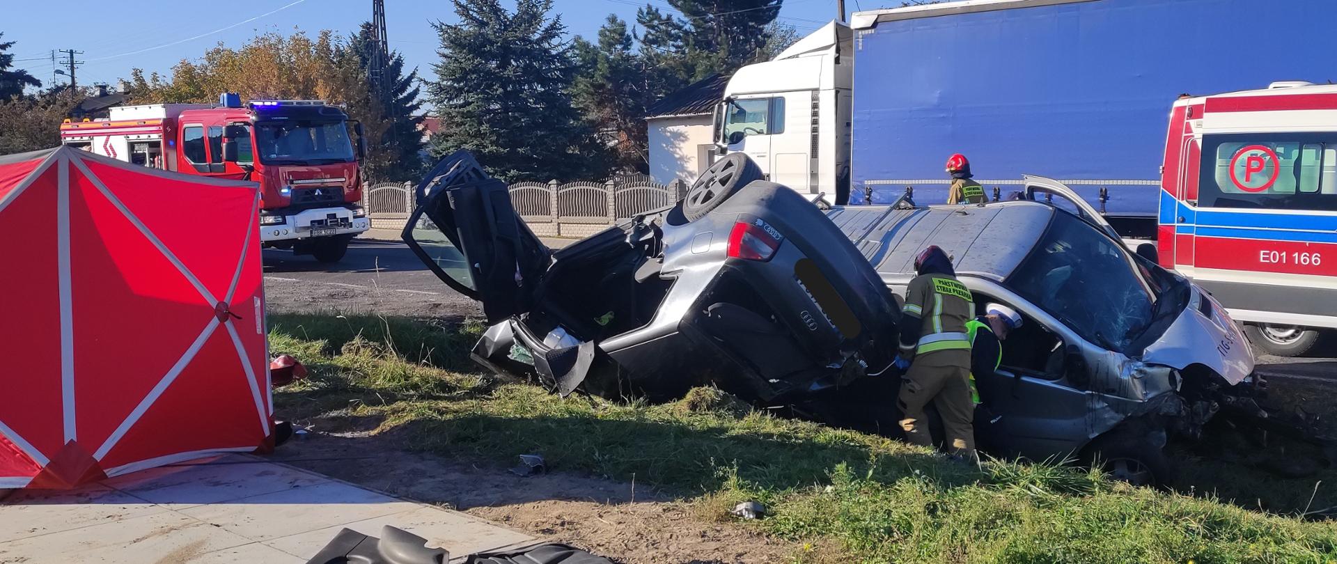 Wypadek drogowy na drodze krajowej 72 w miejscowości Przecław, zderzenie samochodu osobowego z busem, auta znajdują się w rowie, auto osobowe w pozycji na dachu, z licznymi uszkodzeniami, bus uszkodzenia przedniej prawej strony pojazdu, strażak i policjant wykonują czynności ratownicze przy tych pojazdach, na drodze za nimi widać samochód strażacki i karetkę, na poboczu stoi rozłożony czerwony parawan, obok leżą zdemontowane elementy pojazdu.