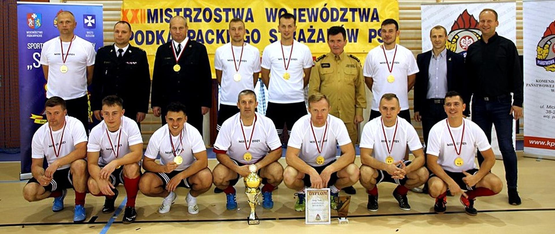 Zdjęcie jest zrobione w hali sportowej. Na zdjęciu reprezentacja Komendy Powiatowej PSP w Leżajsku - zwycięzcy turnieju. Siedmiu mężczyzn, w ubraniach sportowych, z medalami, kuca. Przed nimi stoi puchar oraz dyplom. Za sportowcami stoi dziewięciu mężczyzn. Trzech z nich w mundurach funkcjonariuszy PSP. Z tyłu widać roll-upy oraz baner z napisem XXII Mistrzostwa Województwa Podkarpackiego Strażaków PSP. 