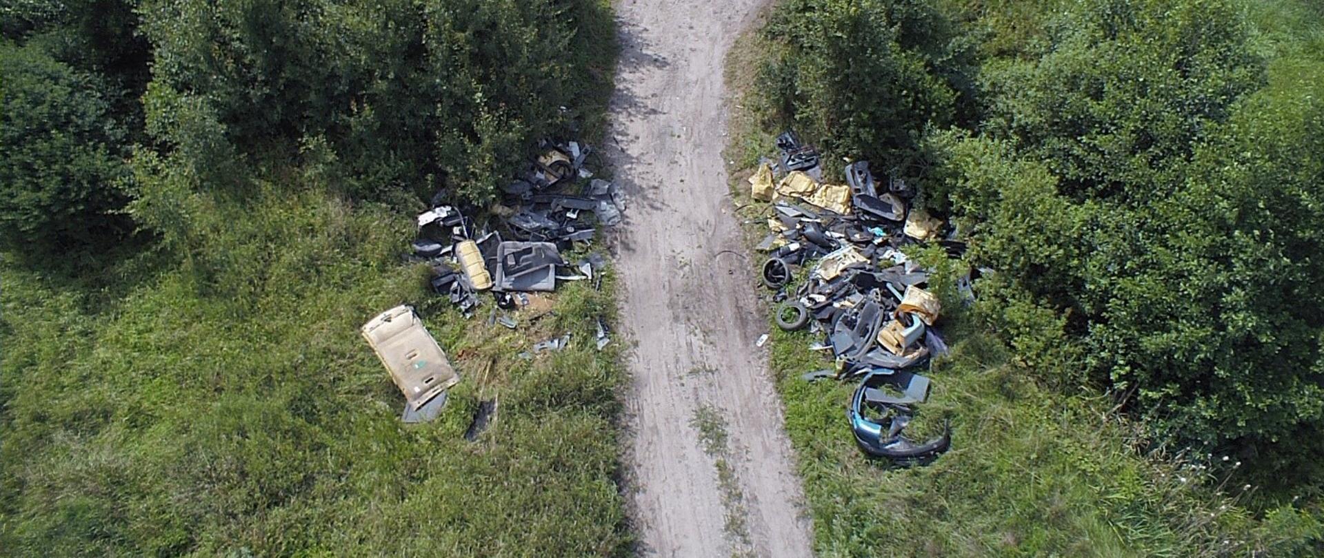 Zdjęcie przedstawia drogę nieutwardzoną oraz teren porośnięty wielogatunkową roślinnością. Hałdy zawierają tworzywa sztuczne oraz elementy wyposażenia wnętrz samochodów. Odpady znajdują się na terenie porośniętym przez wielogatunkową roślinność, zaraz przy drodze nieutwardzonej.