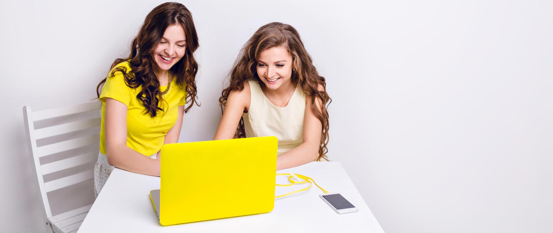 Dwie dziewczyny siedzą za laptopem w żółtej obudowie. Na stole leży smartfon ładowany za pomocą żółtego kabla. Obie dziewczyny uśmiechają się i patrzą na ekran. Jedna ma na sobie żółtą koszulkę, druga - sukienkę.