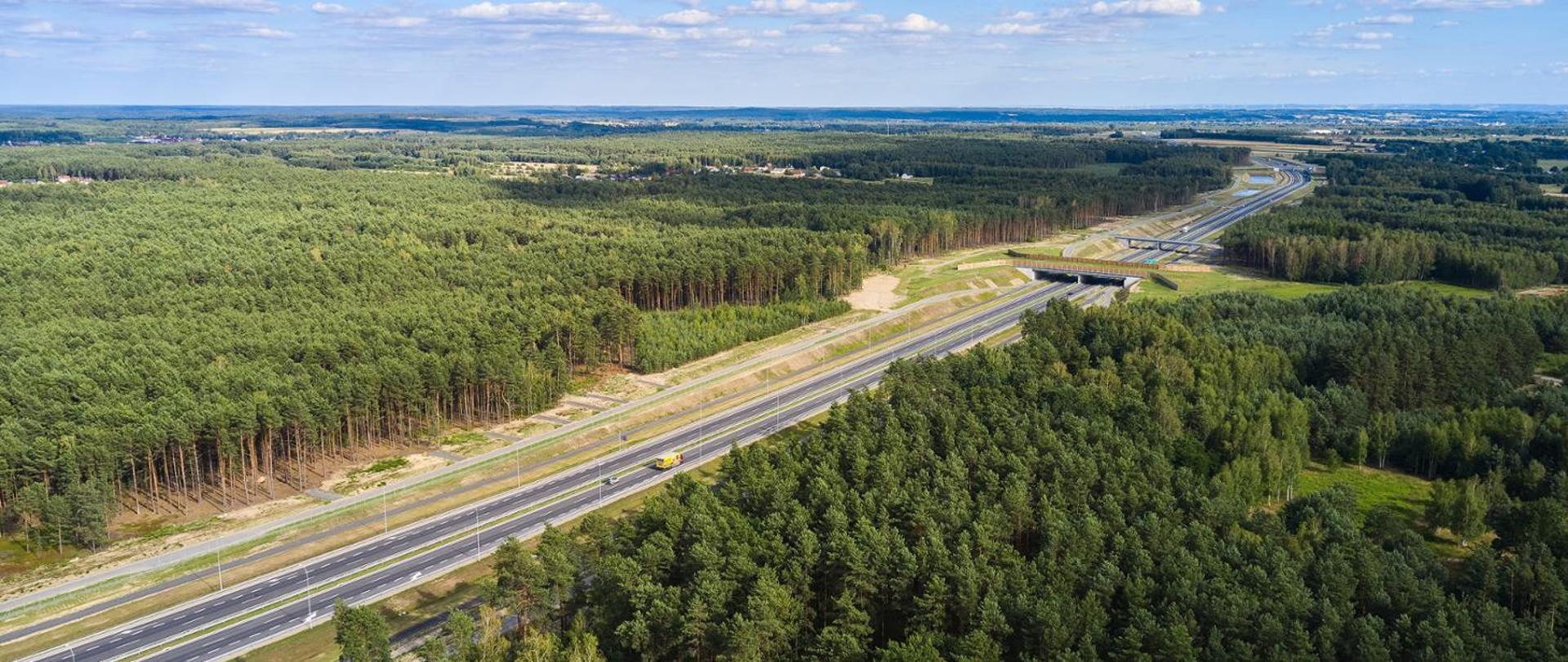 Słoneczny dzień. Widok z powietrza na odcinek drogi ekspresowej przebiegający wśród drzew. Widoczne pojazdy jadące drogą i w oddali przejścia dla zwierząt.