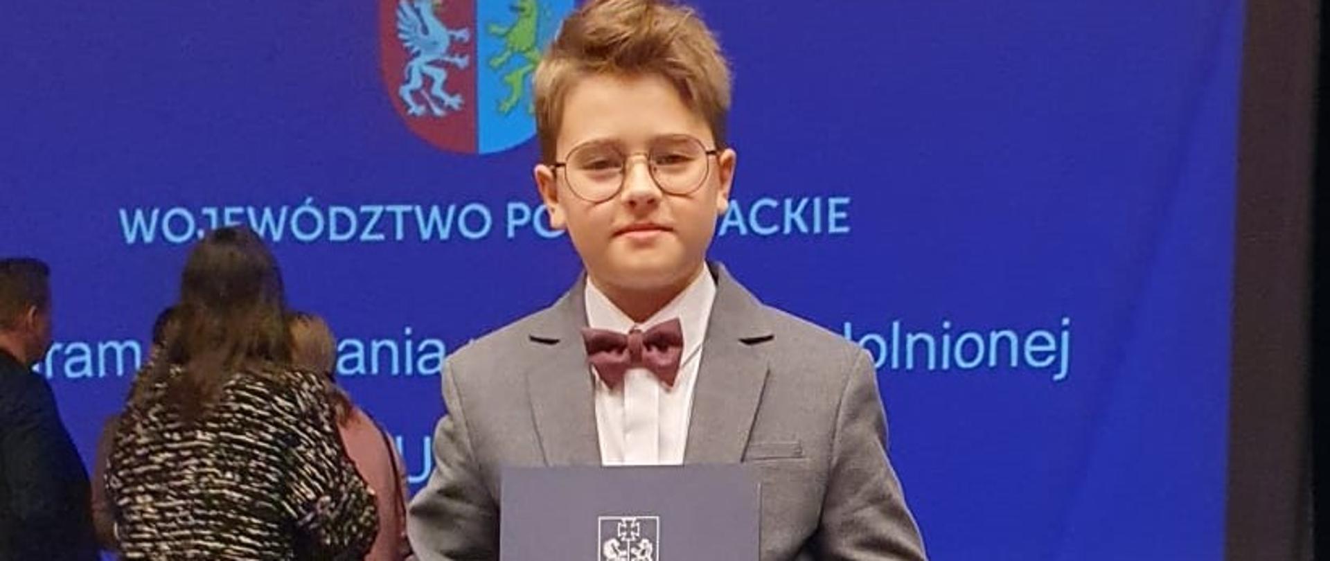 Na zdjęciu laureat stypendium wraz z nagrodą. Na dole, po prawej i lewej stronie zdjęcia widoczne kwiaty w kolorze białym, czerwonym oraz niebieskim.