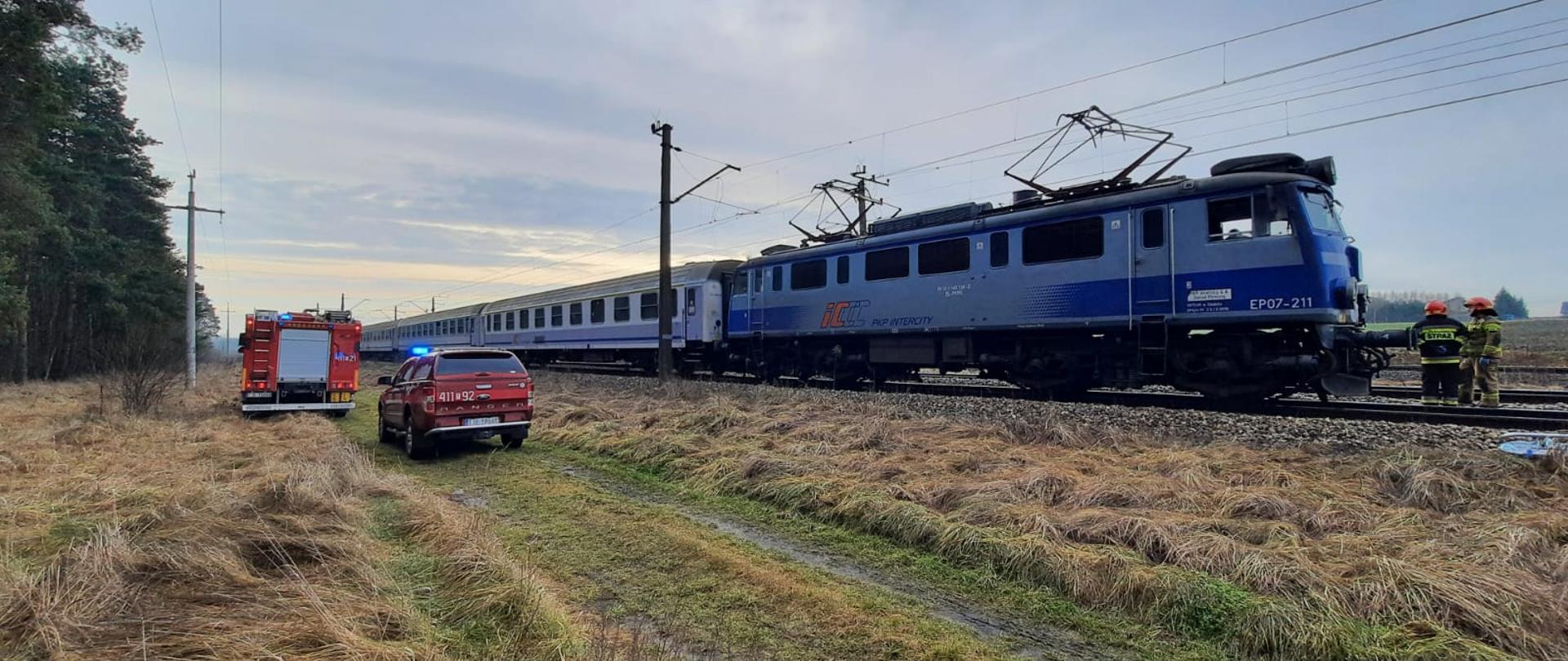 Na zdjęciu pociąg pasażerski TLK na torach kolejowych, samochód ratowniczo-gaśniczy oraz samochód operacyjny zaparkowane na polanie obok torów kolejowych.