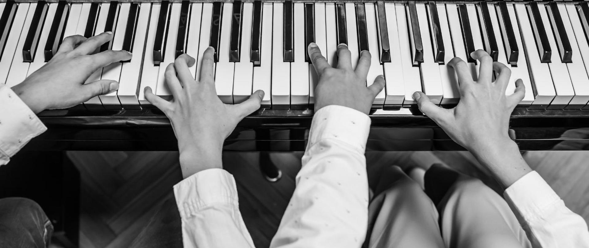 czarno-białe zdjęcie rąk dwóch osób grających na fortepianie, perspektywa z lotu ptaka, dłonie odbijają się w desce fortepianu